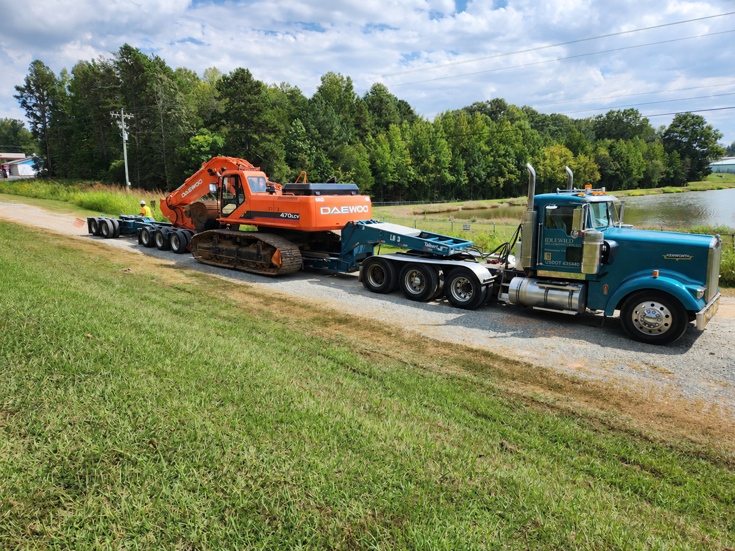 Blue truck and trailer carrying an orange excavator with a worker beside the trailer