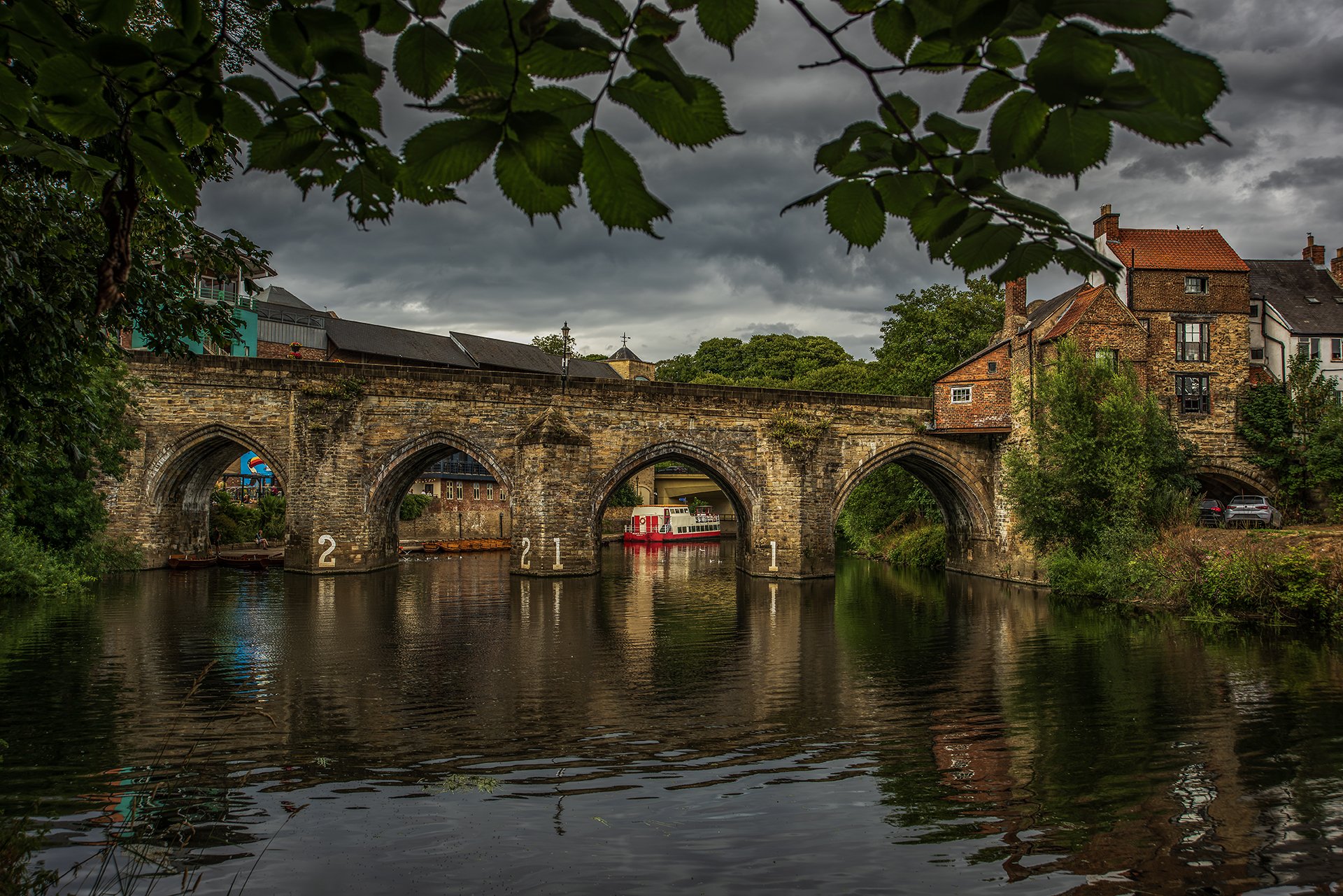 Stone bridge and red-and-white boat reflected in a river beneath leaves.