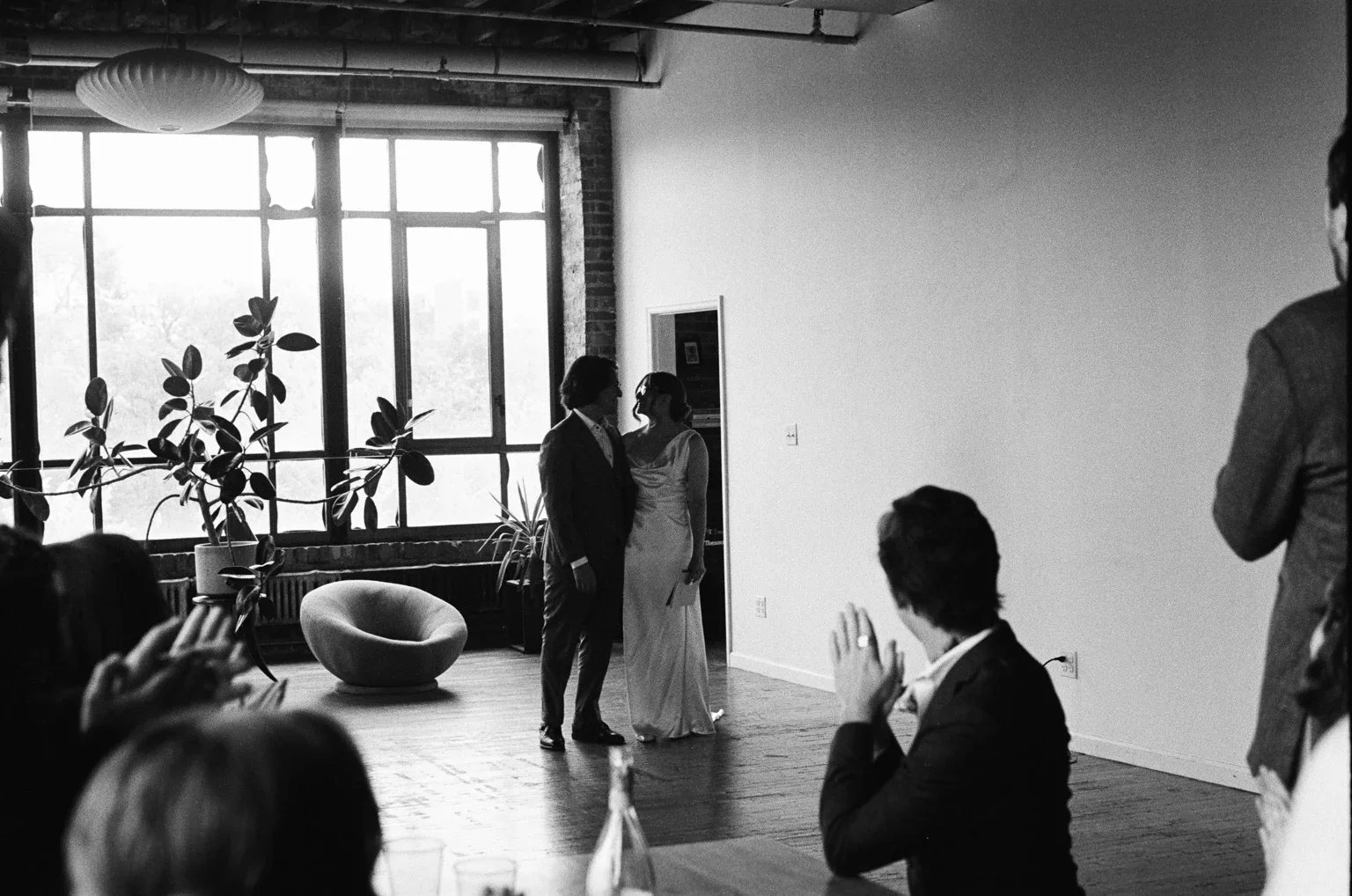 Guests watching a wedding speech in a sunlit room, photographed in black and white