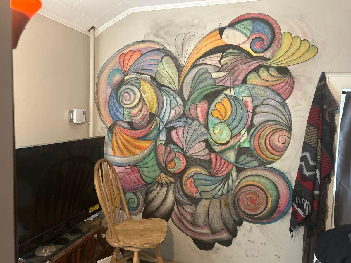 Original room photograph of a colorful abstract wall mural, partially obscured by a television and wooden chair