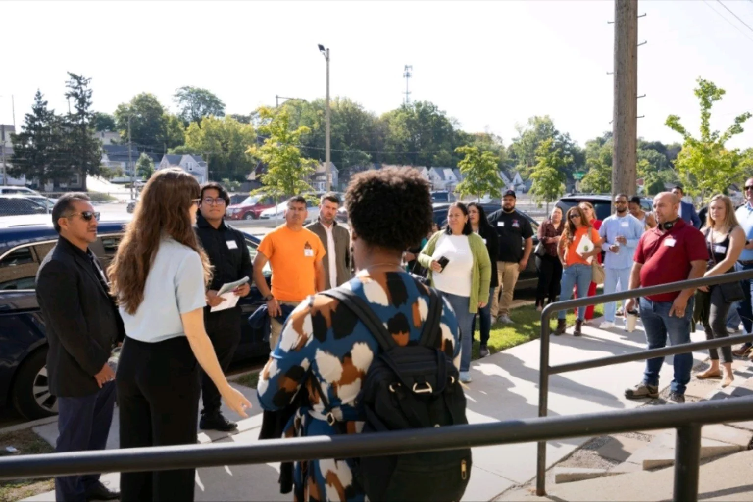 A group gathered outdoors in a neighborhood, listening together beside a sidewalk and nearby homes.