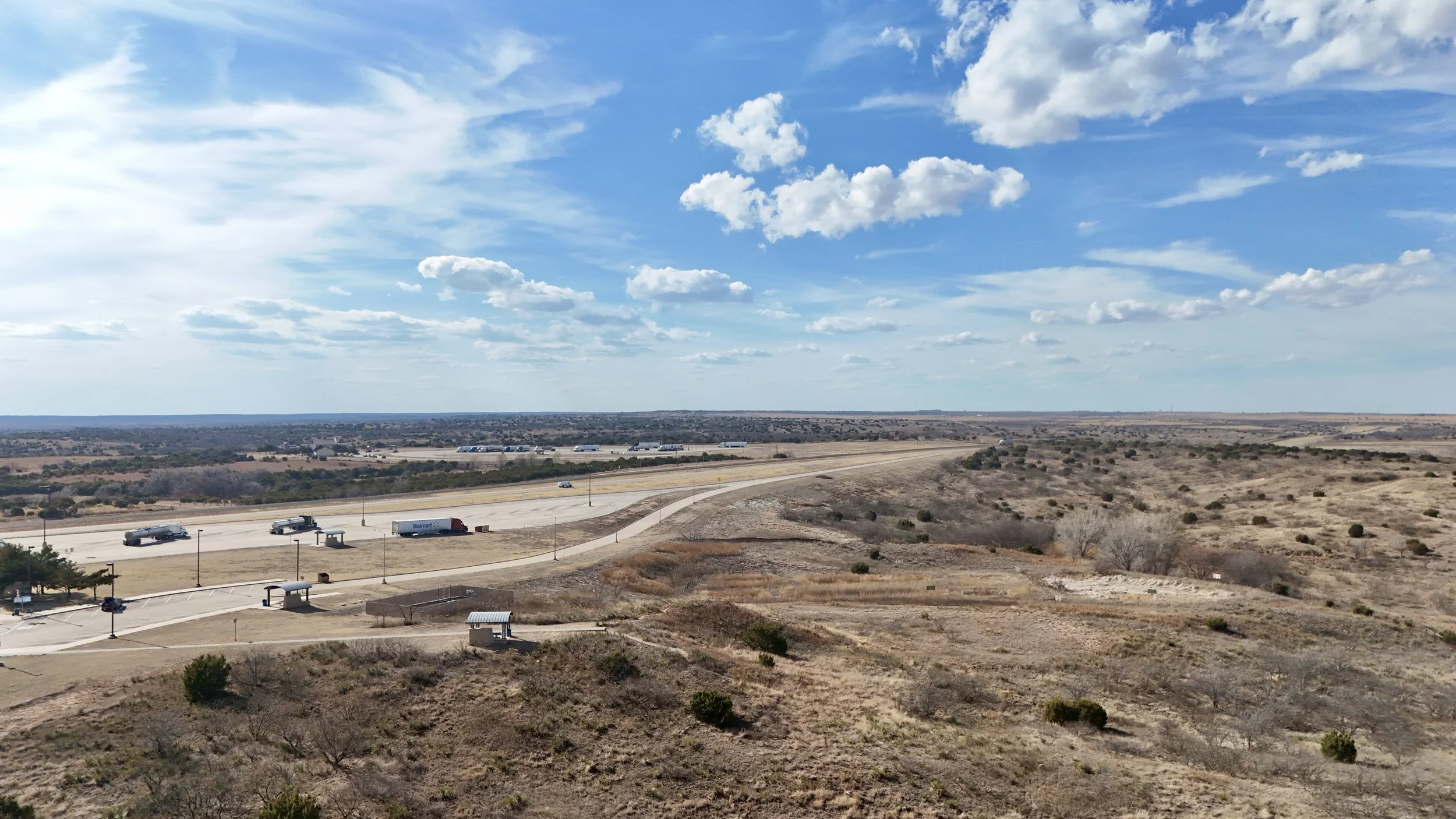Wide aerial view of open land, access roads and the surrounding landscape beneath a bright sky.