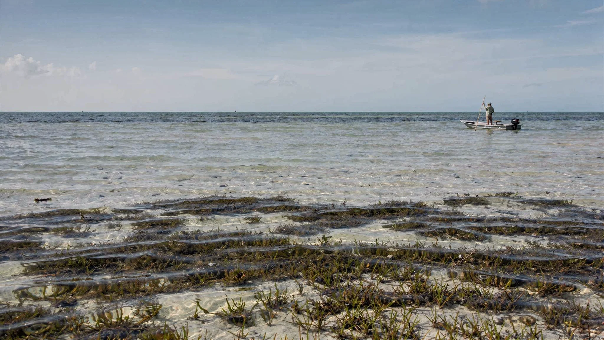 Wide panoramic view of a Florida Keys bonefish flat at low tide &mdash; a guide poles a skiff in the distance, turtle grass in the foreground, white sand bottom beneath clear water.