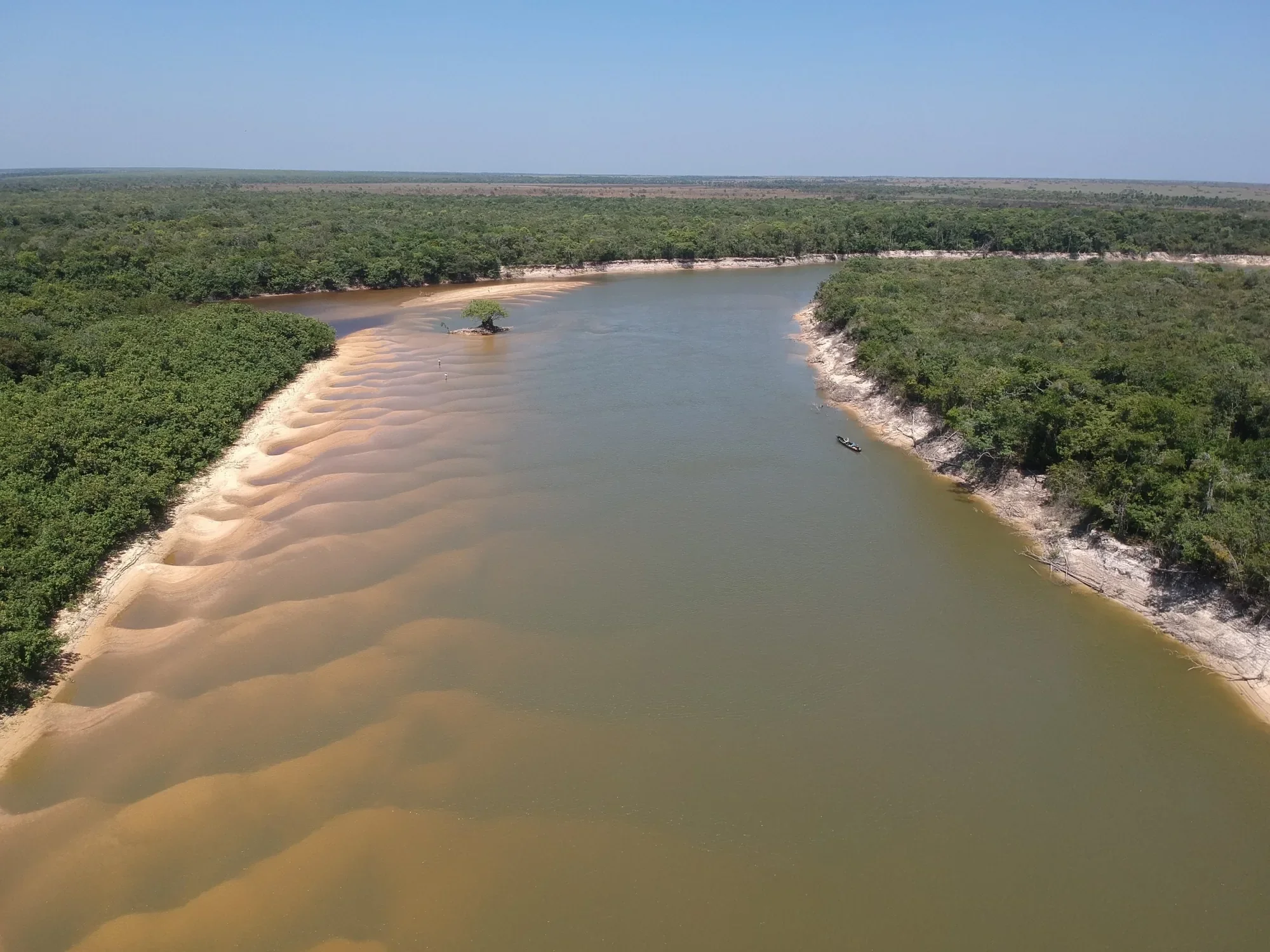 Aerial view of a wide, shallow river in the dry season: rippled sand visible beneath the water, pale eroded banks standing above it, gallery forest on both sides and a small boat near the right bank