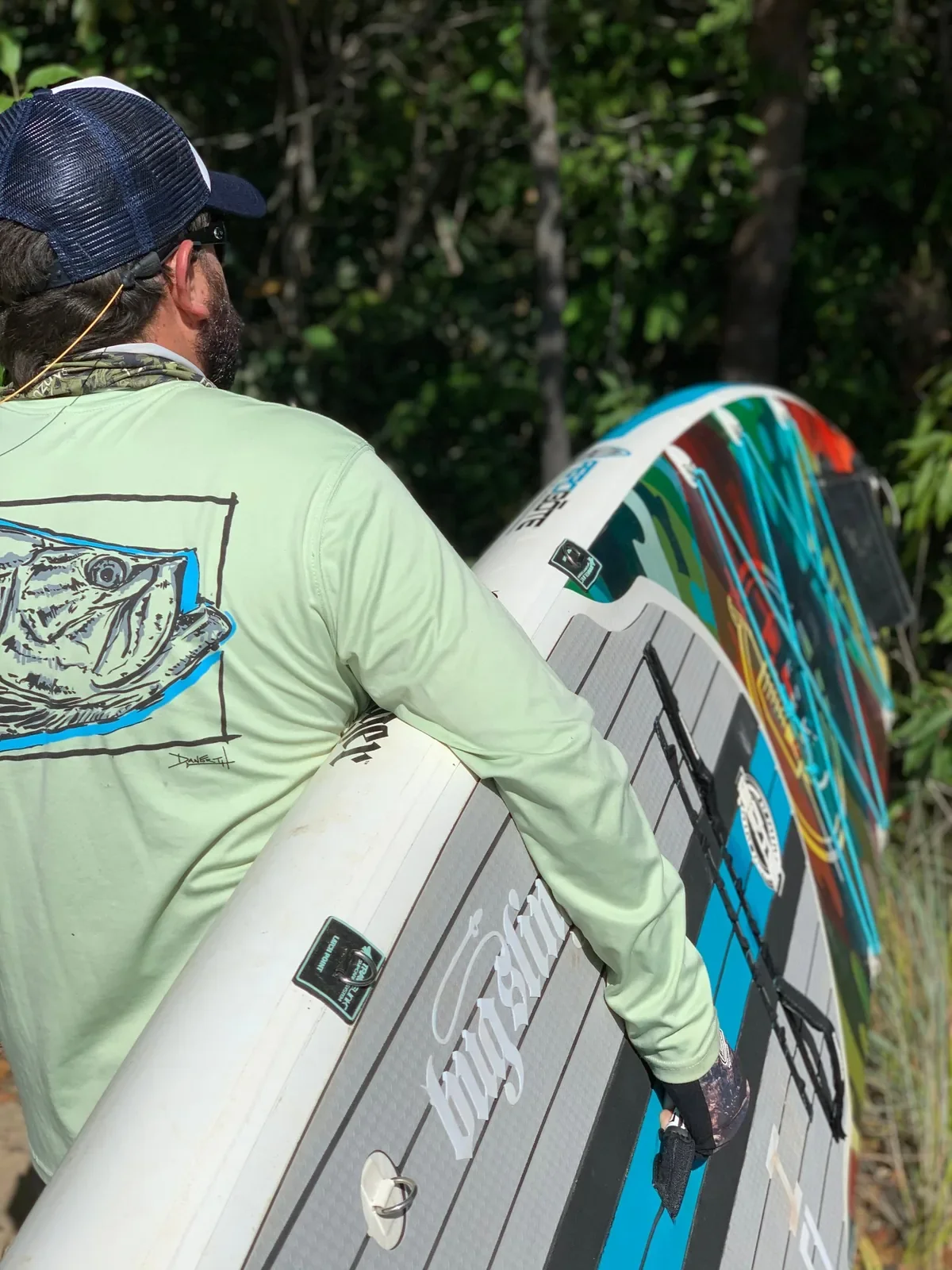 Angler in a sun shirt carrying an inflatable paddle board along a forest trail toward a lagoon
