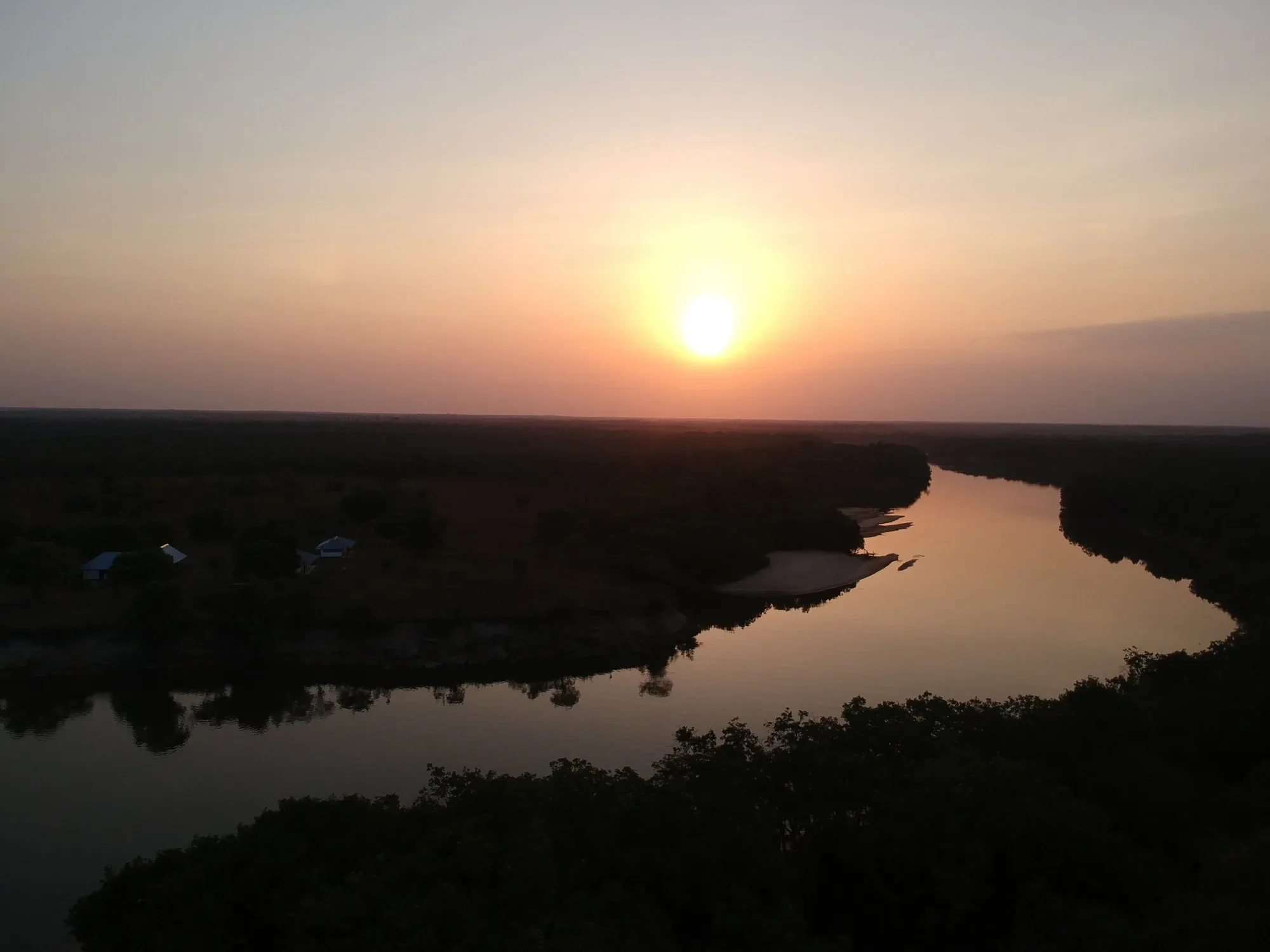 Aerial view of a river bend at sunset, a riverside lodge on the left bank and savanna forest to the horizon
