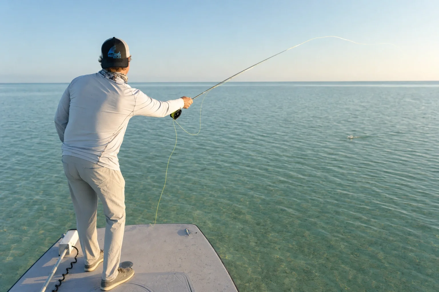 Fly angler casting from the bow of a flats skiff at dawn, chartreuse fly line forming a loop in the air above clear teal water.