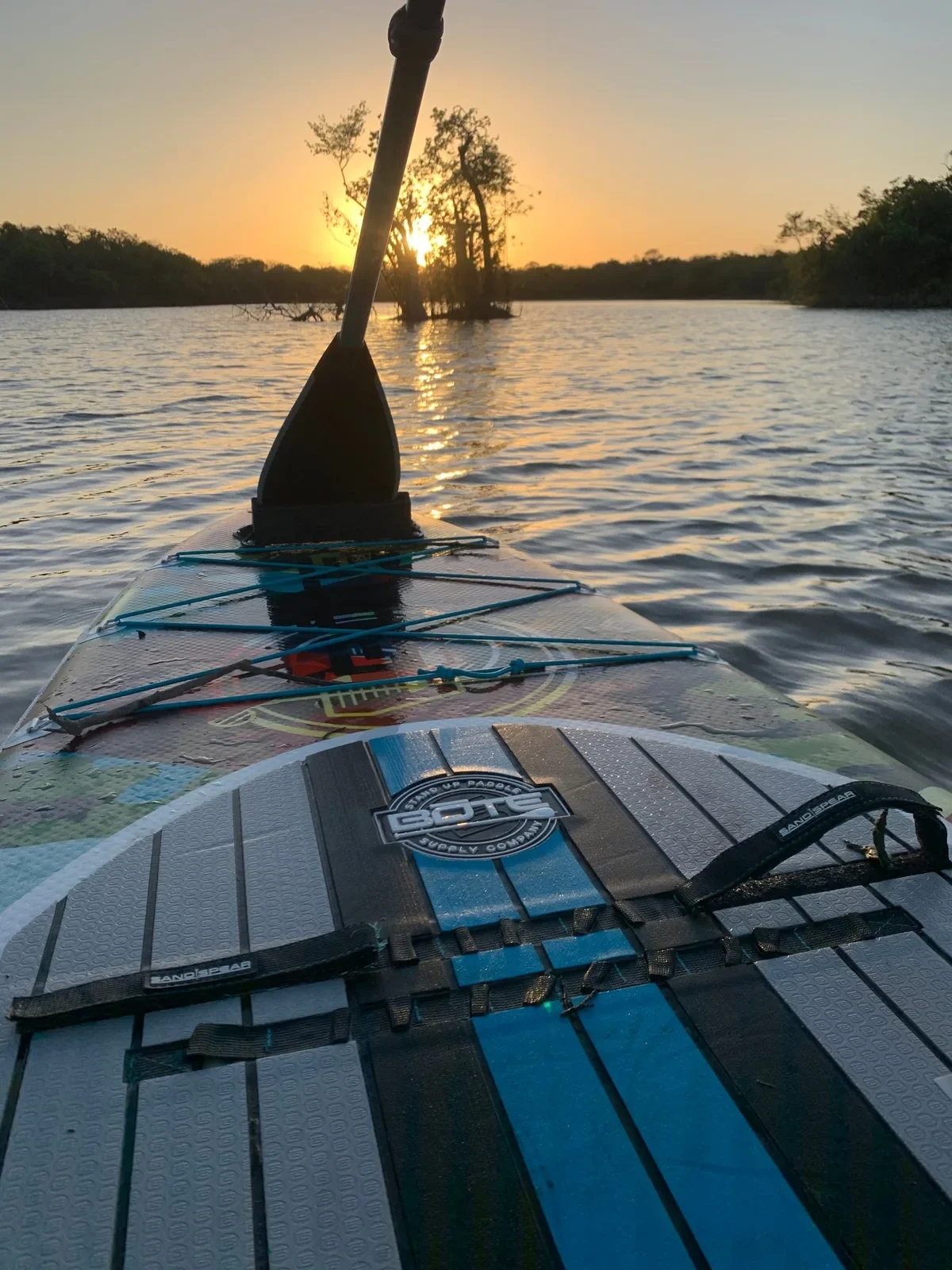 View from the deck of a paddle board across a calm lagoon at sunset, paddle blade in the foreground