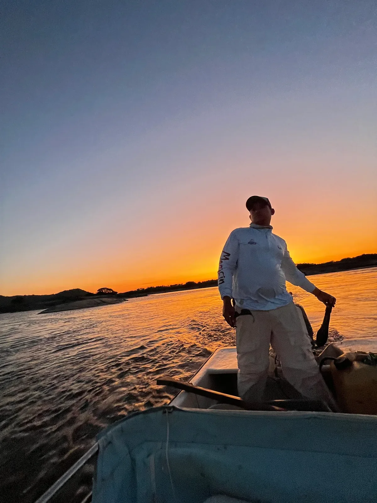 Fishing guide Gustavo Novoa standing at the tiller of a river boat at sunset, granite outcrops on the far bank
