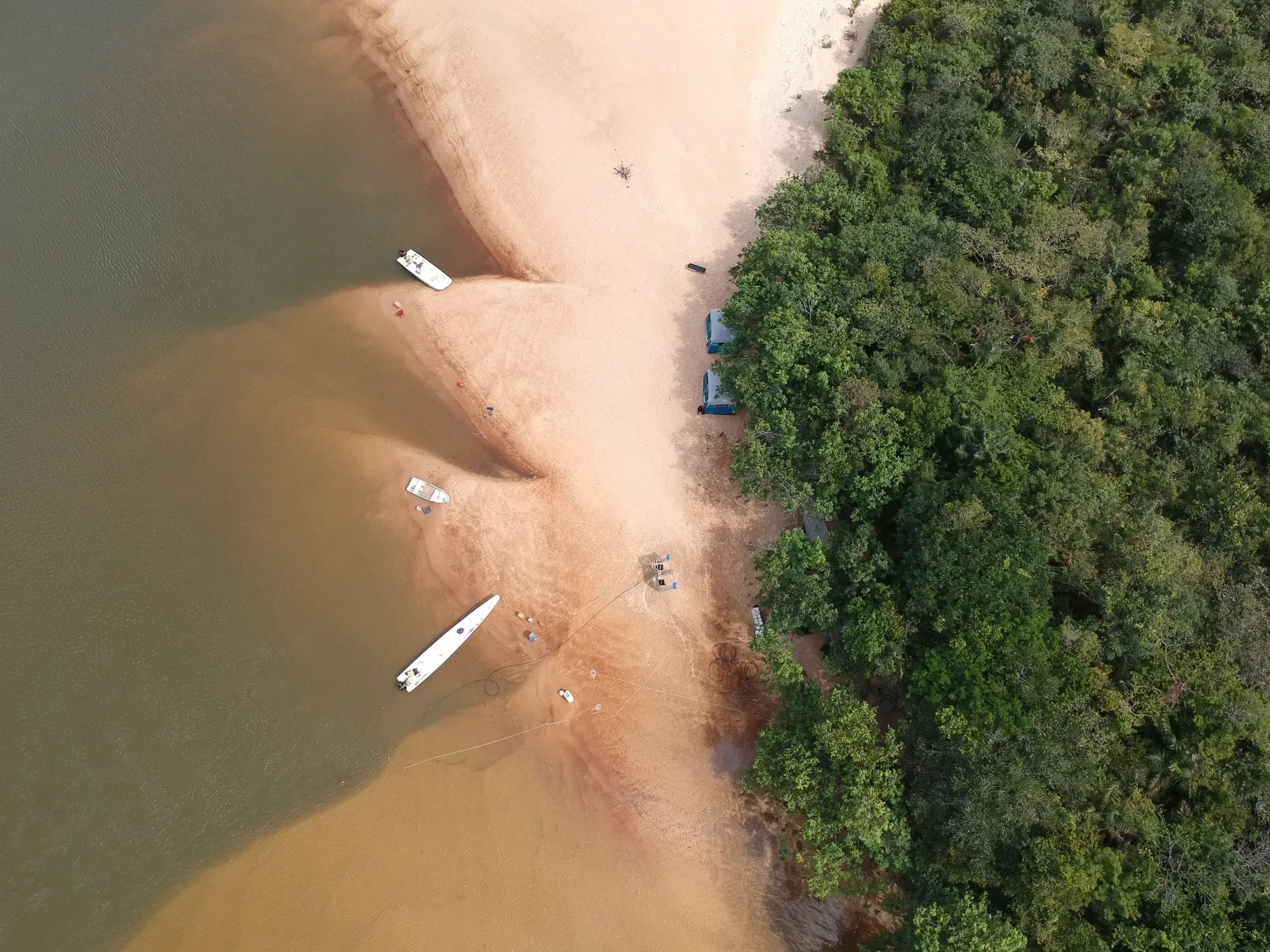 Overhead view of a riverside fishing camp: boats pulled onto a pale sandbar, tents tucked under the forest edge