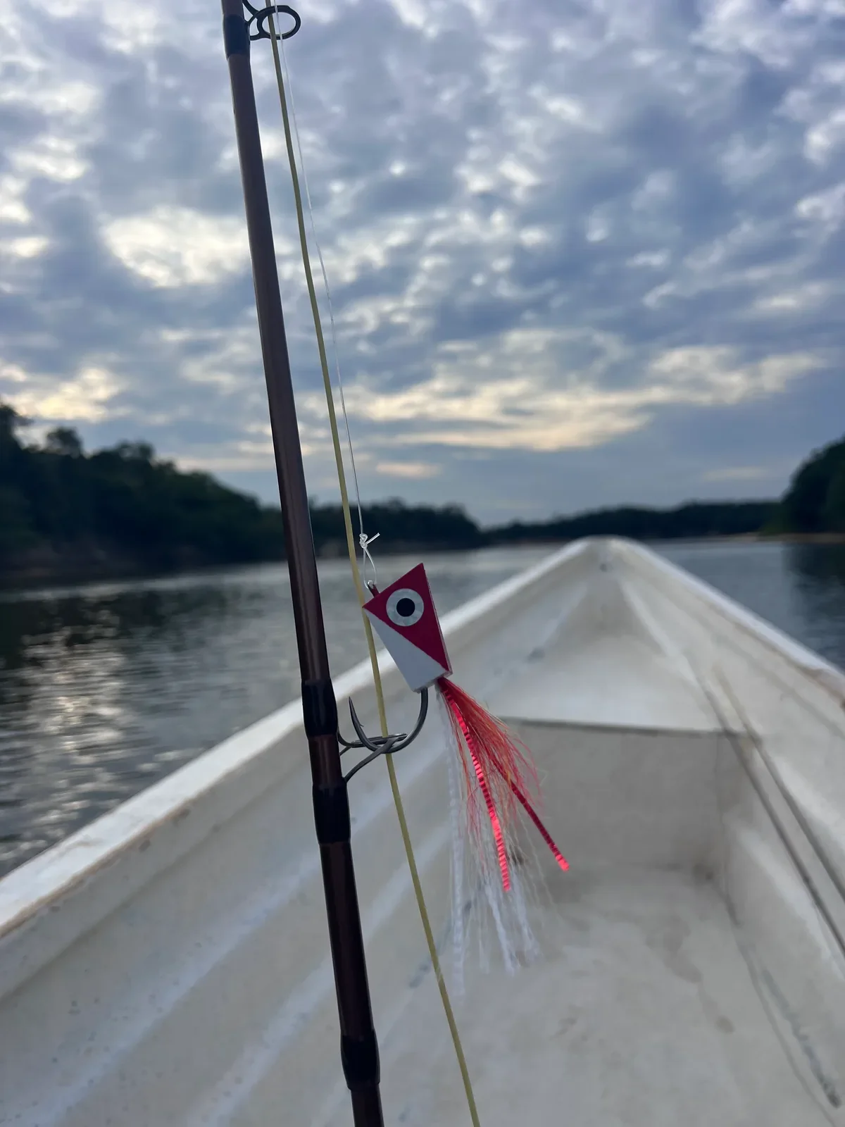 Red and white foam-headed popper fly hanging from a fly rod in the bow of a river boat under a cloudy evening sky
