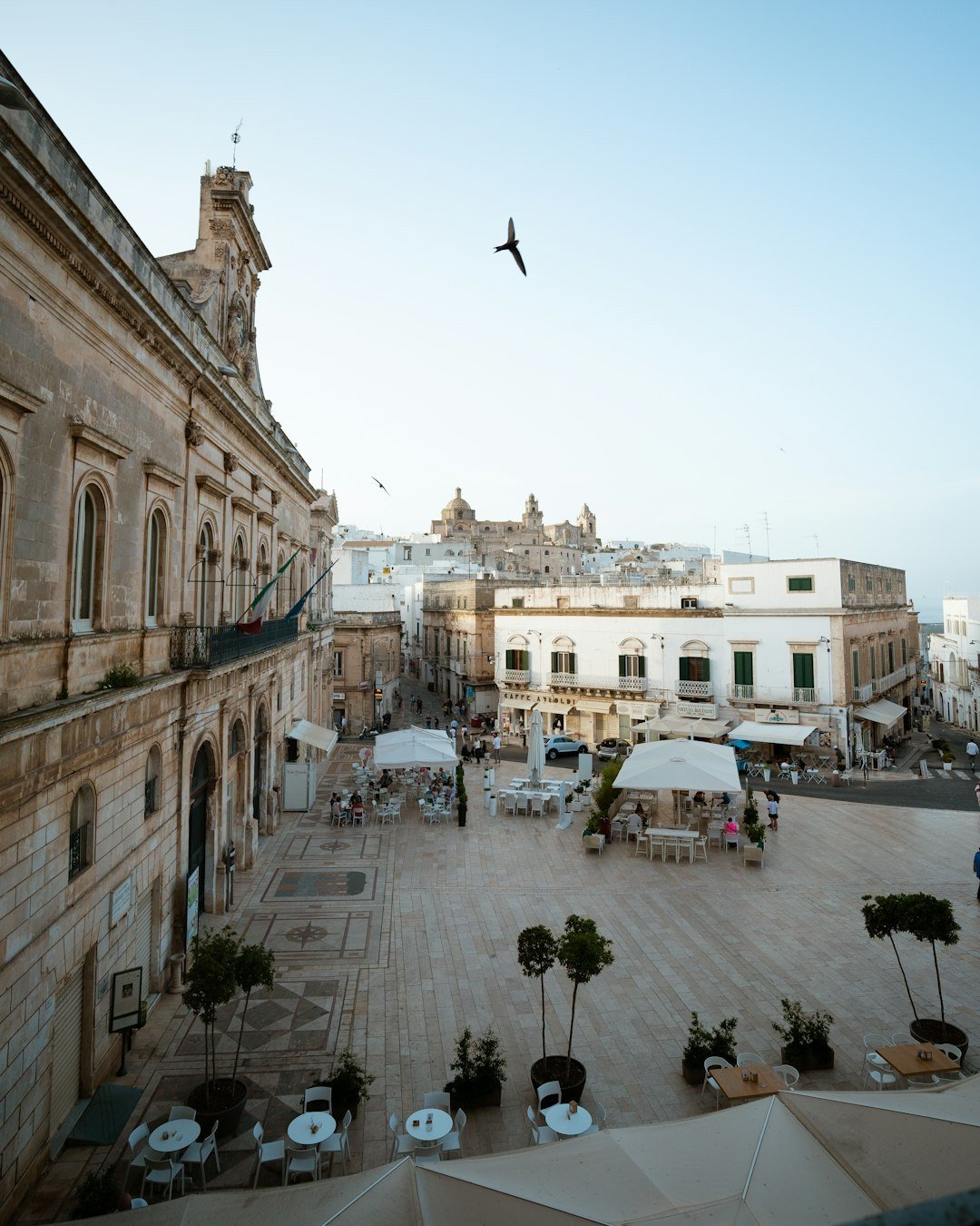 Cafe tables and people in an Ostuni piazza surrounded by pale stone buildings.