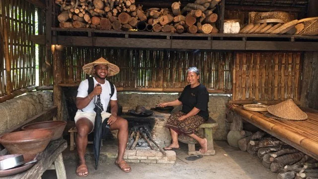 Mike Randle in a straw hat, sitting in a rustic open-air hut beside a woman cooking over a wood fire