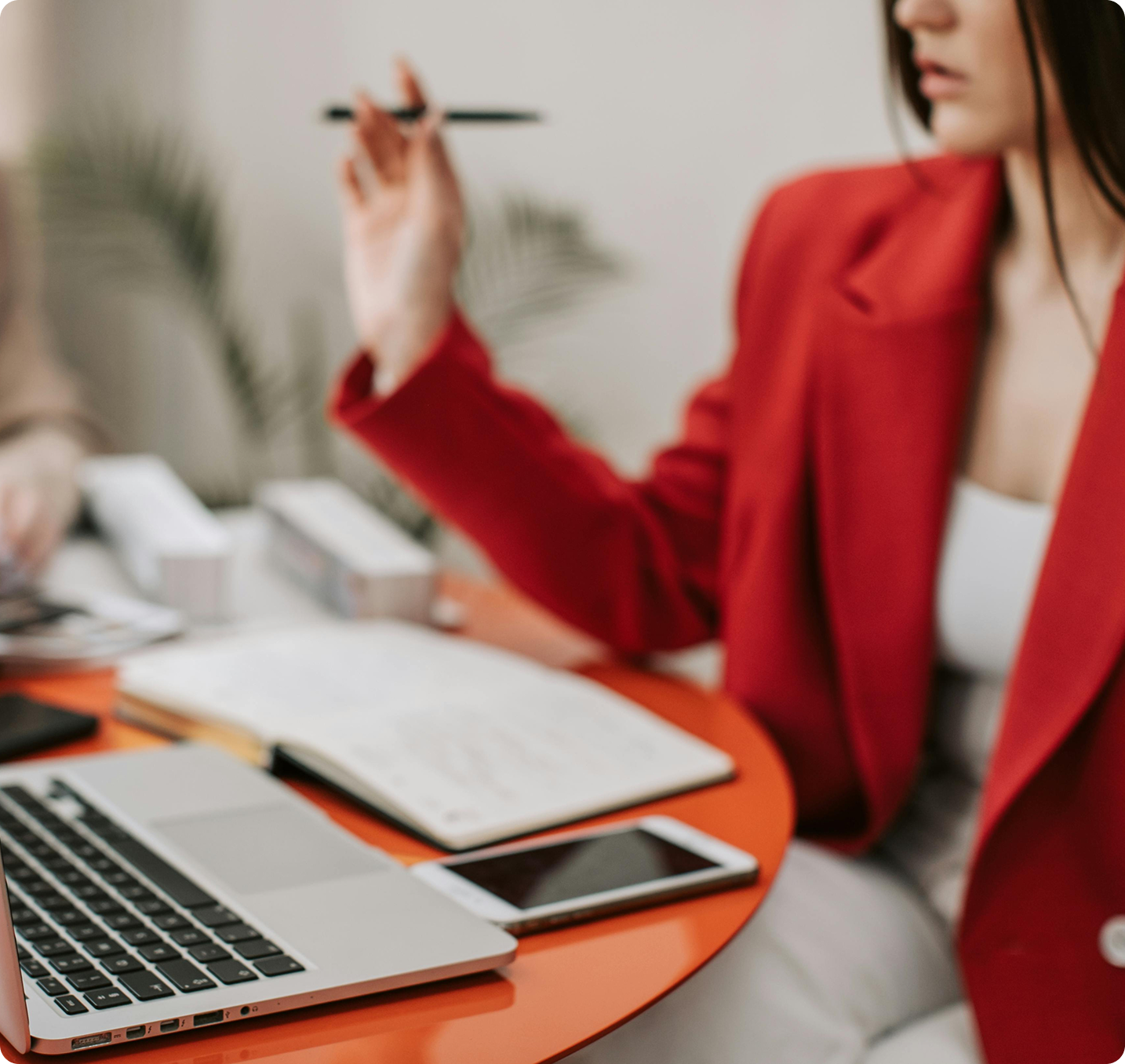 Person in a red blazer gesturing with a pen while speaking at a desk.