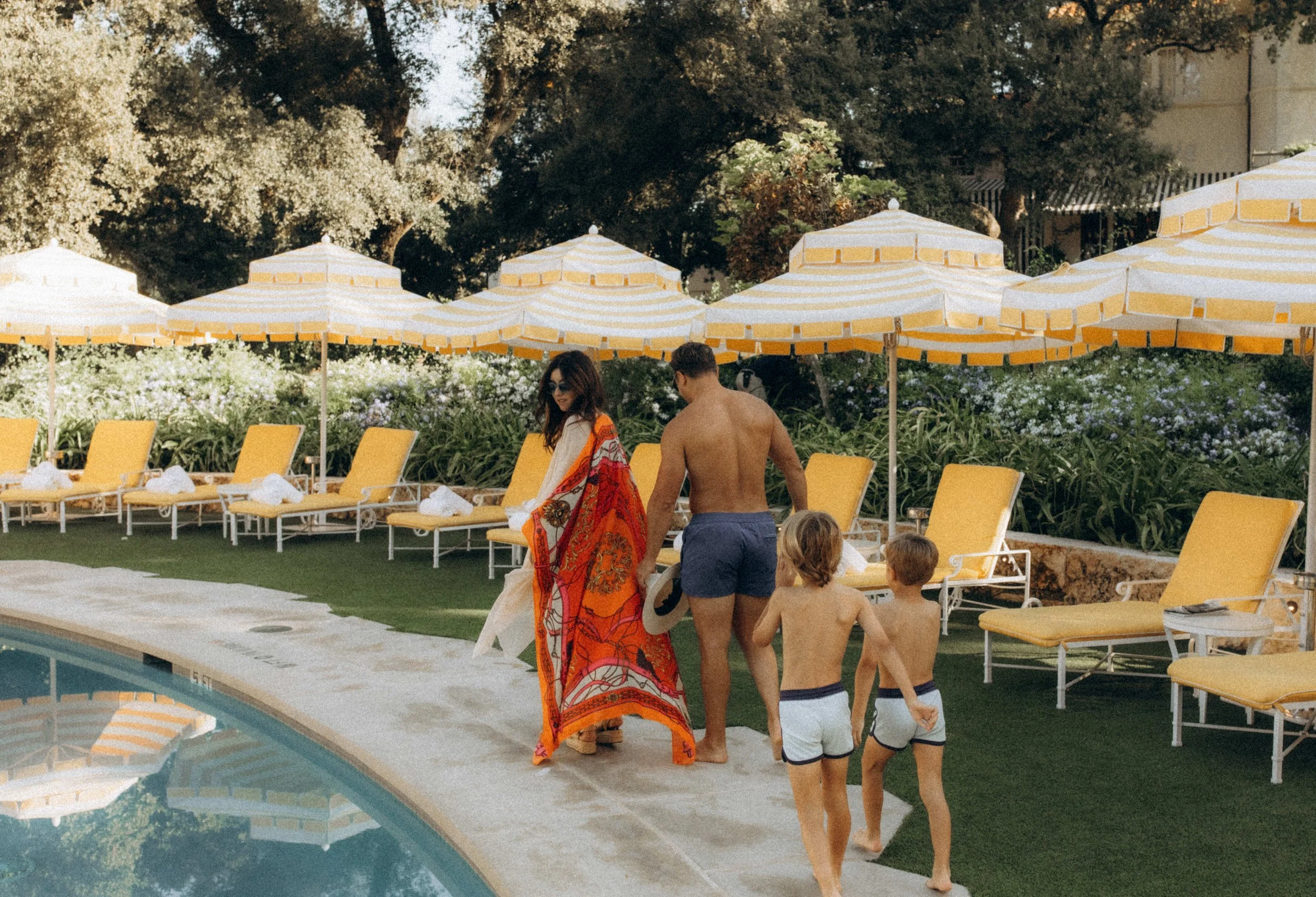 A family walking by a pool, mom in a bright orange patterned wrap, dad and two boys in swim shorts, with yellow striped umbrellas and chairs behind them