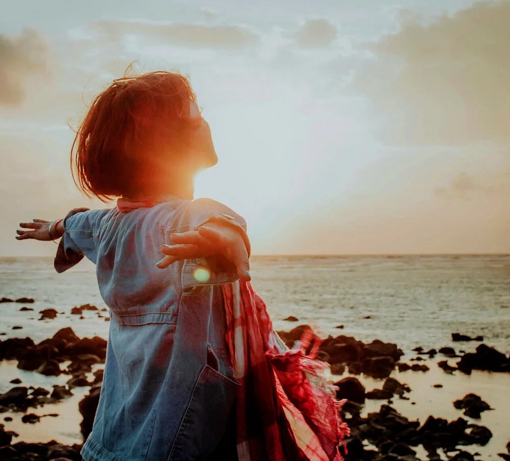 Woman with open arms facing the sunset by the sea, representing the openness of past life regression