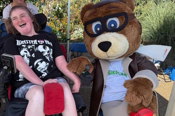 Young participant in a wheelchair smiling with the Forward Facing bear mascot under a canopy