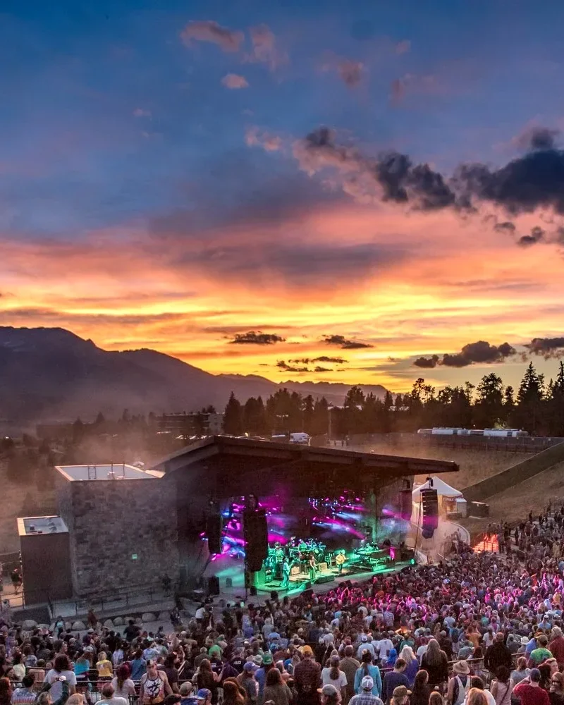 Large crowd enjoying a live concert at Dillon Amphitheater in Colorado, with a brightly lit stage, mountain backdrop, and colorful sunset sky.