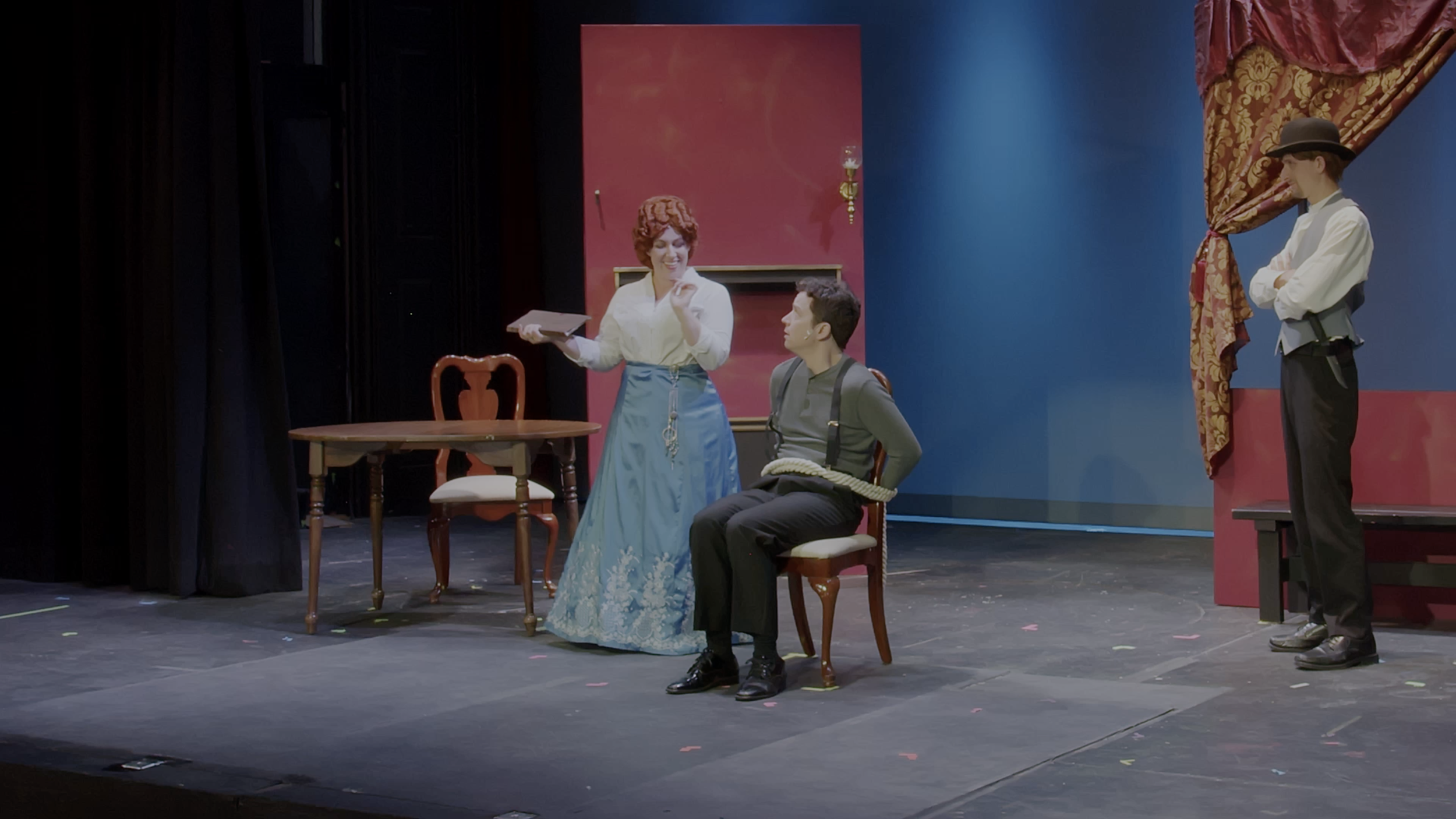 Mrs. Frost smiles down at Nathaniel, who is tied to a chair. A man armed with a knife watches from the side.