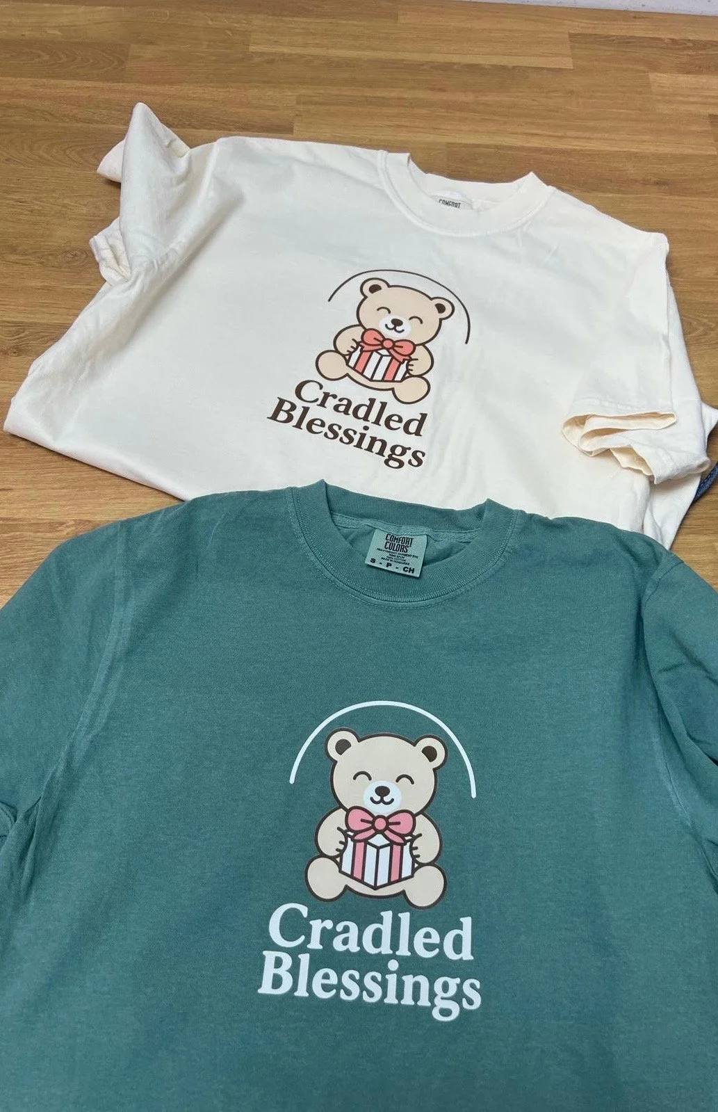 Cream and teal Cradled Blessings chapter T-shirts with the bear logo