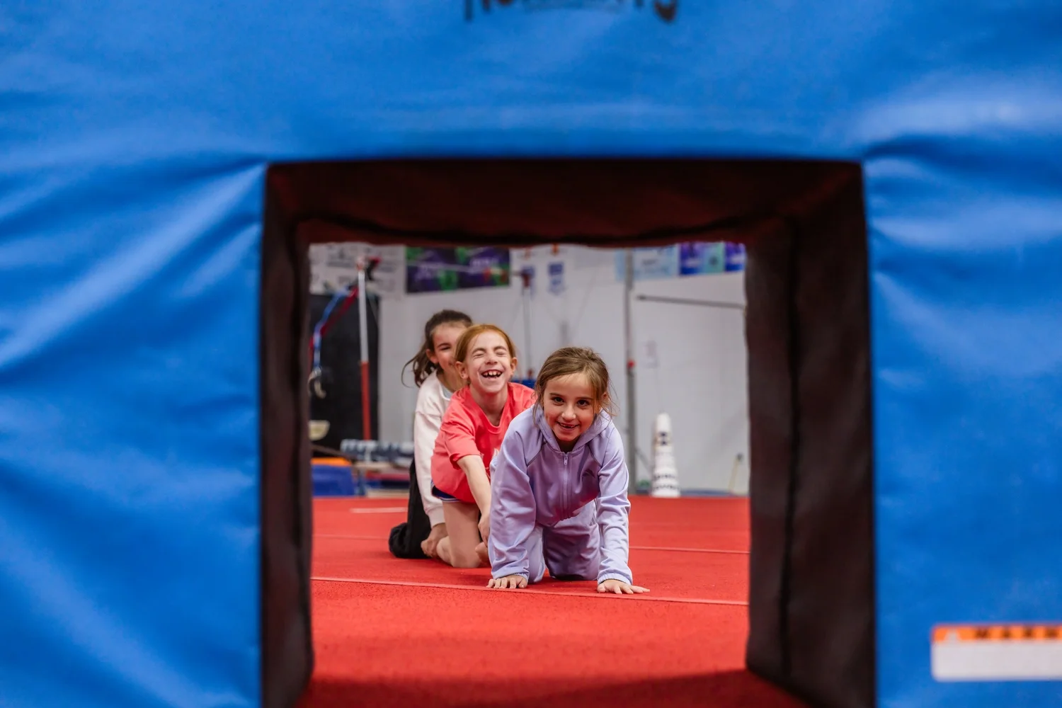 Smiling kids crawl toward the camera through a blue foam tunnel on the gym floor.