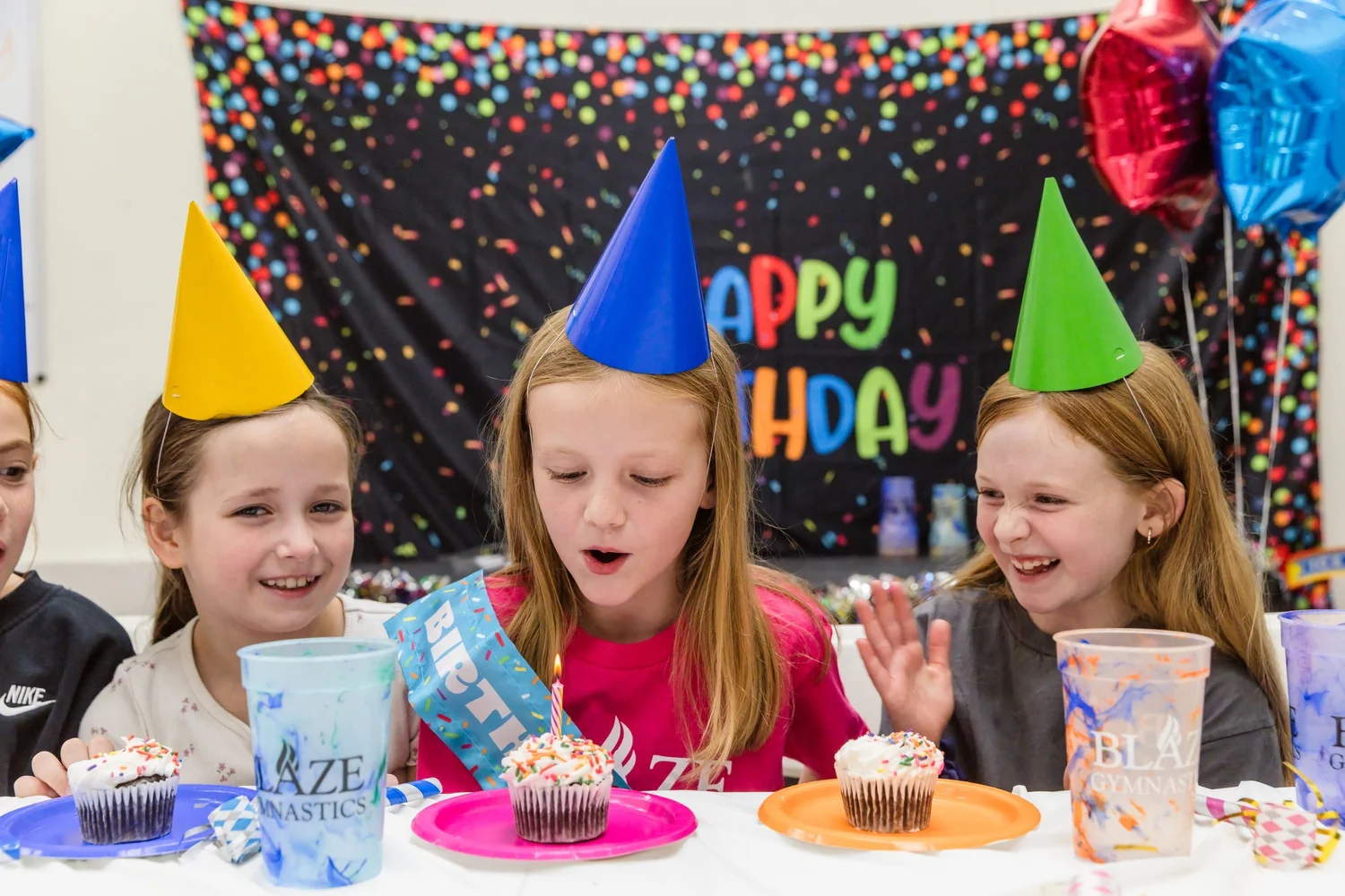 A birthday child in a sash blows out the candle on her cupcake as smiling friends in party hats look on.