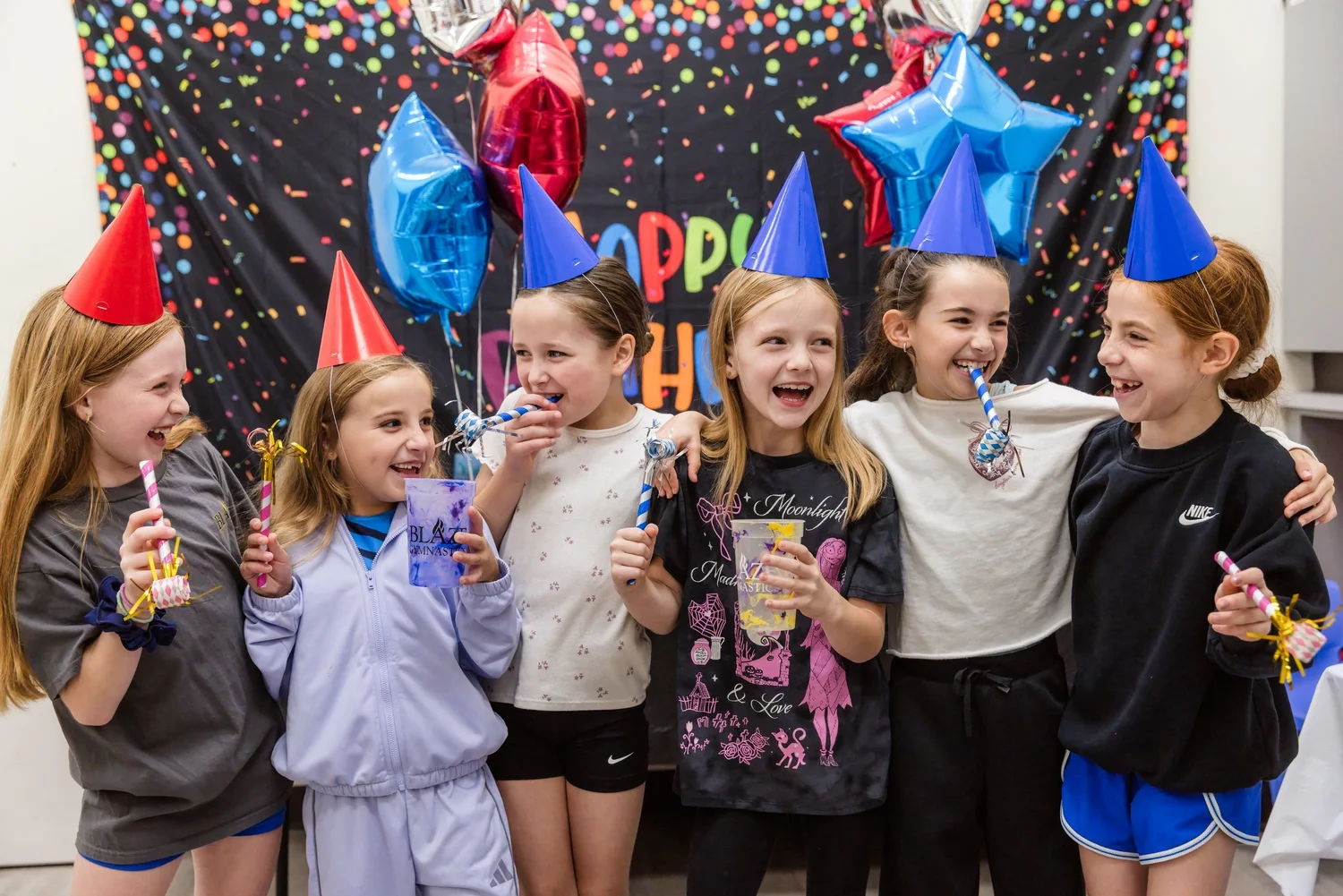 A group of laughing kids in party hats hug and wave noisemakers in front of a Happy Birthday backdrop.