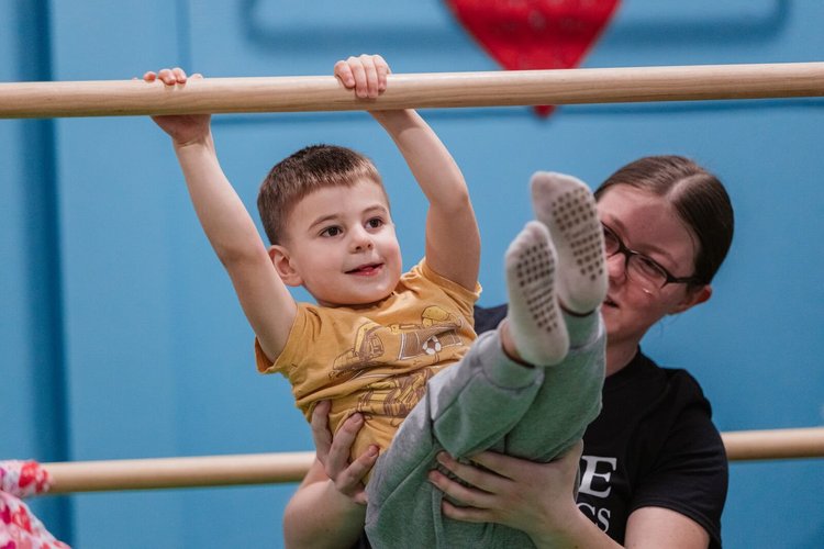Coach supporting a smiling preschool boy as he hangs from a low bar in the tots gym