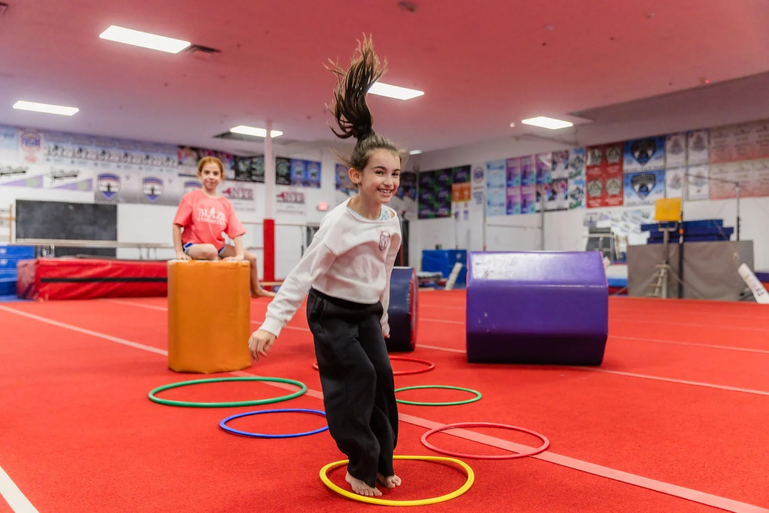 A smiling child jumps into a hoop on the red gym floor at Blaze Gymnastics, with colorful foam shapes and team banners behind.