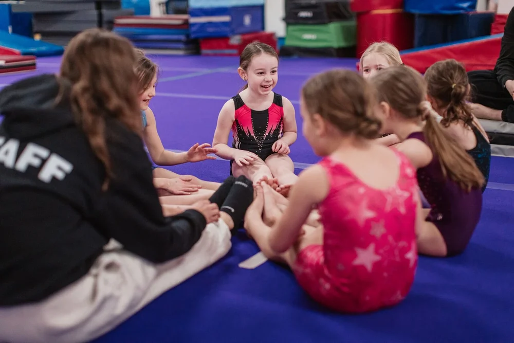 Blaze coach leading a smiling group of young gymnasts in a seated stretching circle