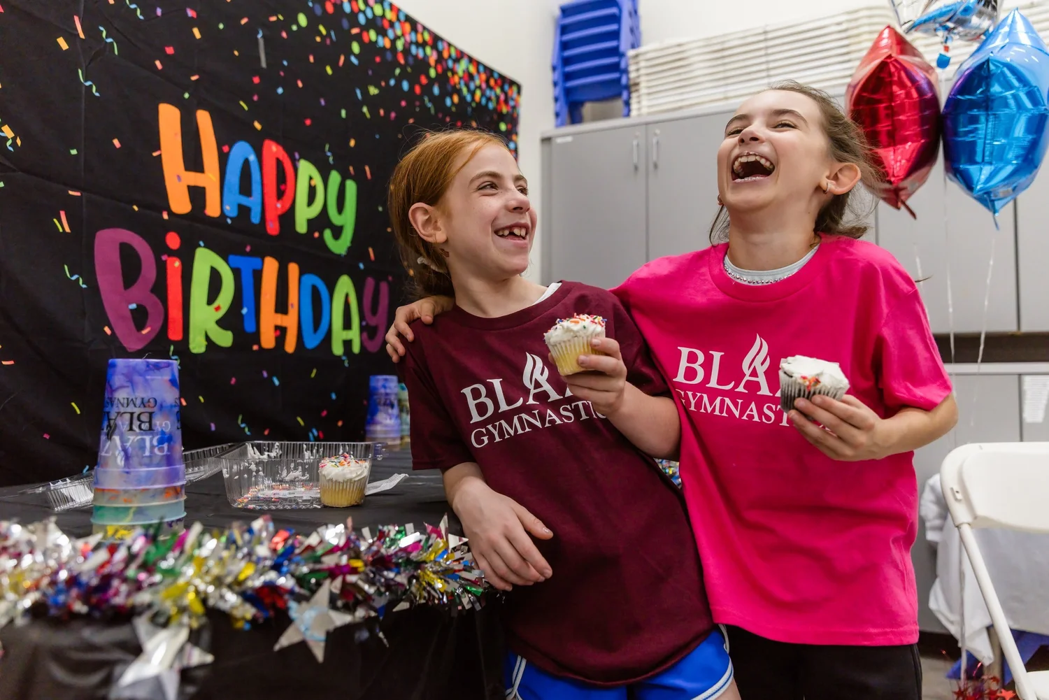 Two friends in Blaze Gymnastics shirts laugh together while holding cupcakes in front of a Happy Birthday backdrop.