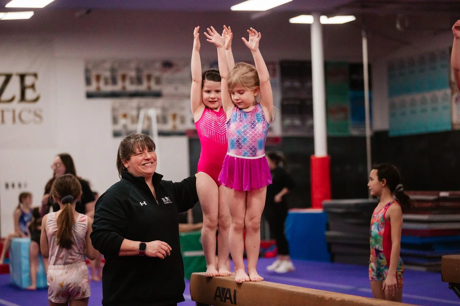 Smiling Blaze Gymnastics coach supporting two young girls standing on a low balance beam with their arms raised