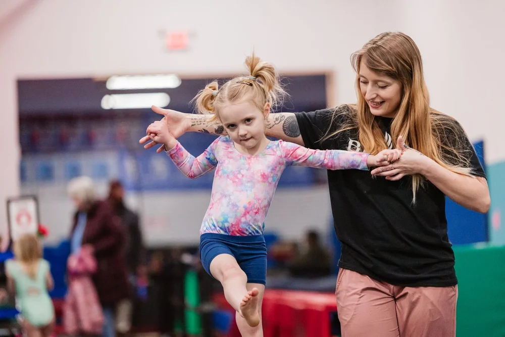 Blaze coach holding a toddler's hands as she steps along a low balance beam in the preschool gym