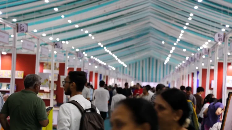 Crowds of readers fill a long aisle of book stalls under a teal and white canopy