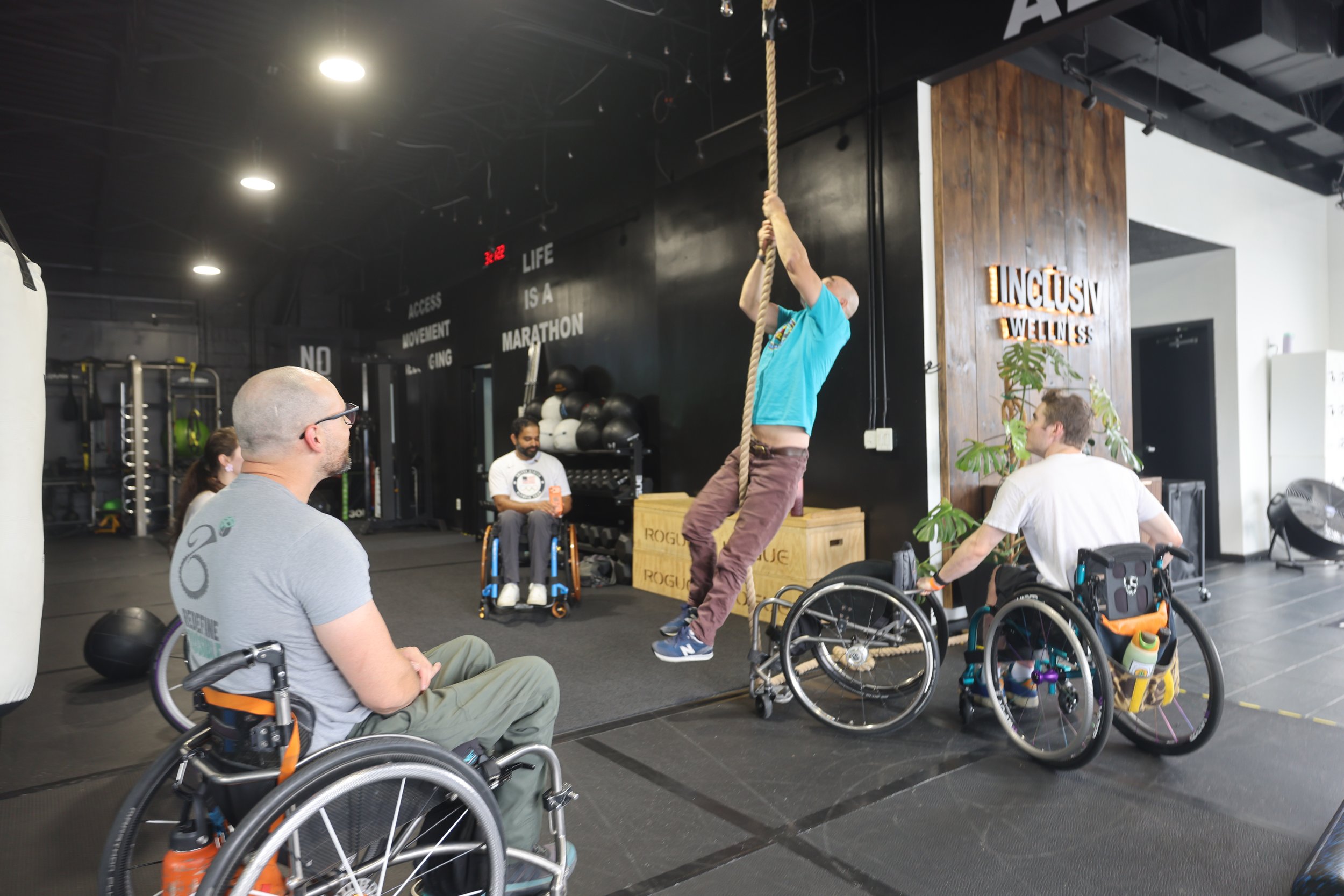 A member climbing the rope while members in wheelchairs cheer him on at INCLŪSIV Wellness