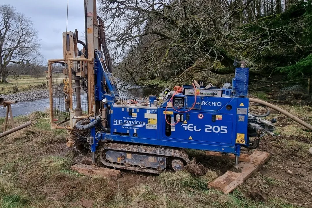 A blue tracked rotary drilling rig conducting geotechnical ground investigation works on a riverbank in Scotland.