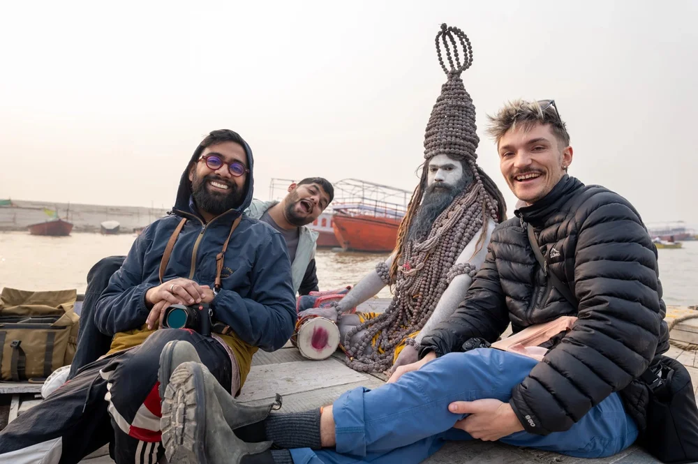 Photographers and a man wearing a beaded headdress sit together beside a river.