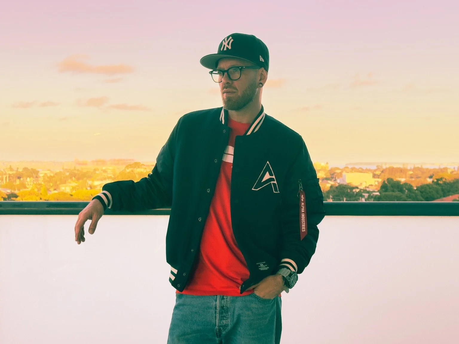A person in glasses, a black cap, a dark jacket and a red shirt leans beside a railing.