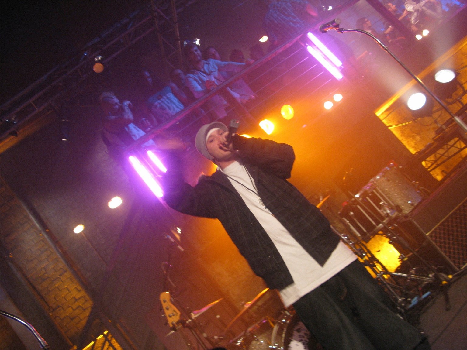 A performer holding a microphone under purple and amber stage lights, with a balcony audience behind.