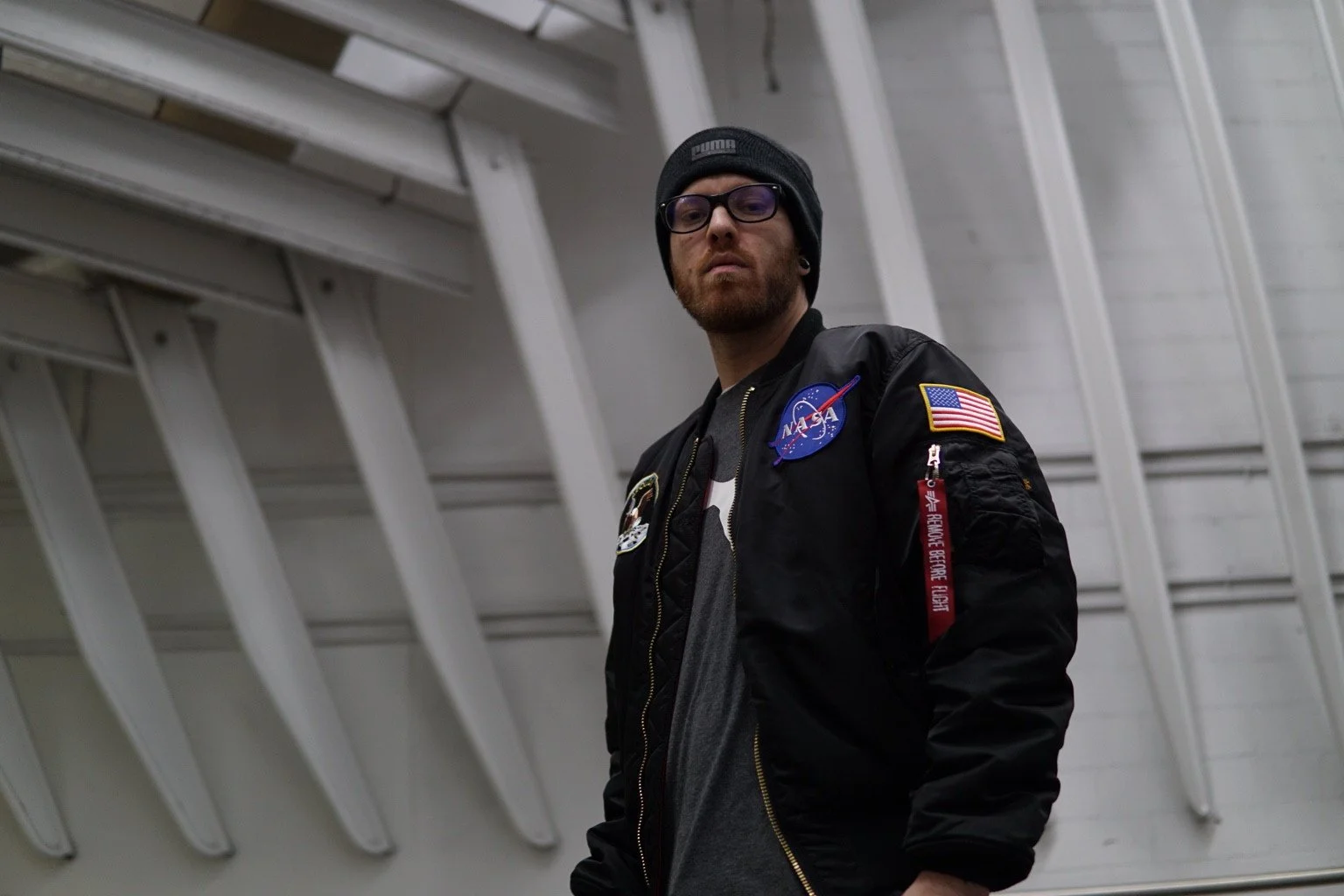 Low-angle portrait of a person wearing glasses, a knit cap and a black bomber jacket.