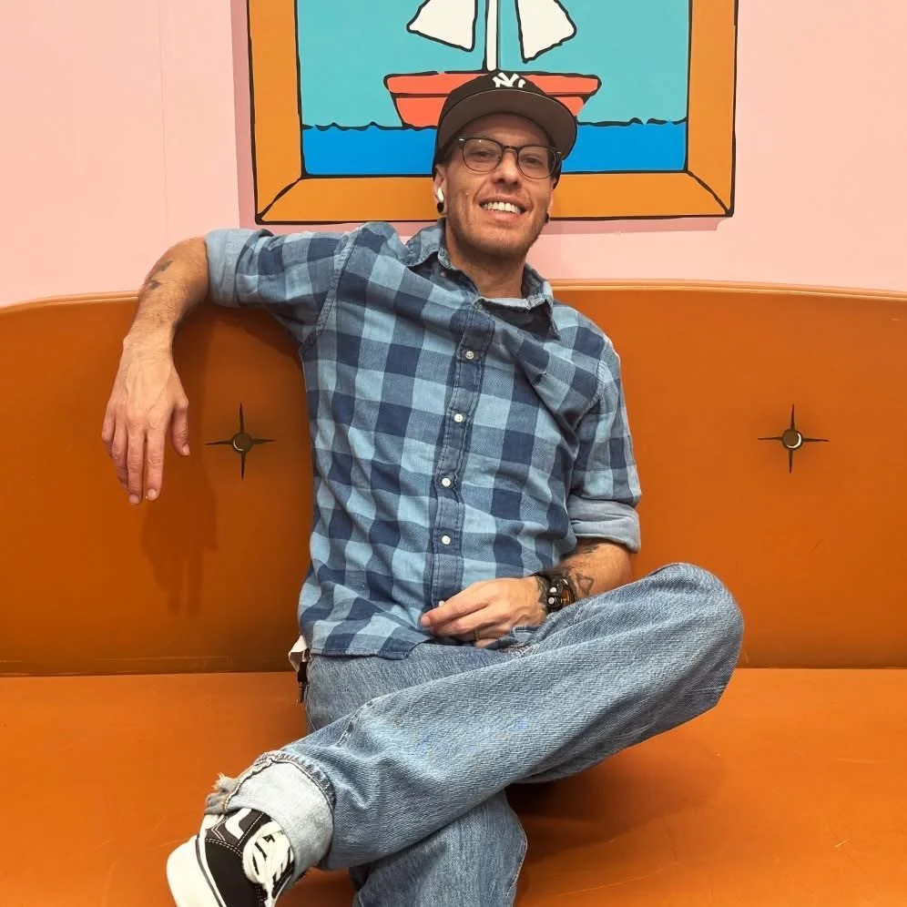 A smiling person in a black cap and blue checked shirt sits cross-legged on an orange sofa beneath a sailboat illustration.