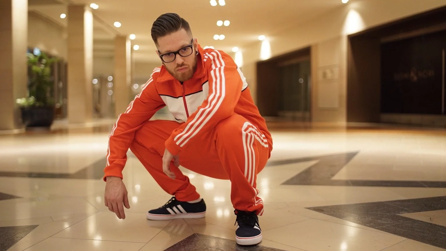 A person in an orange-and-white tracksuit crouches on a patterned floor in a warmly lit lobby.