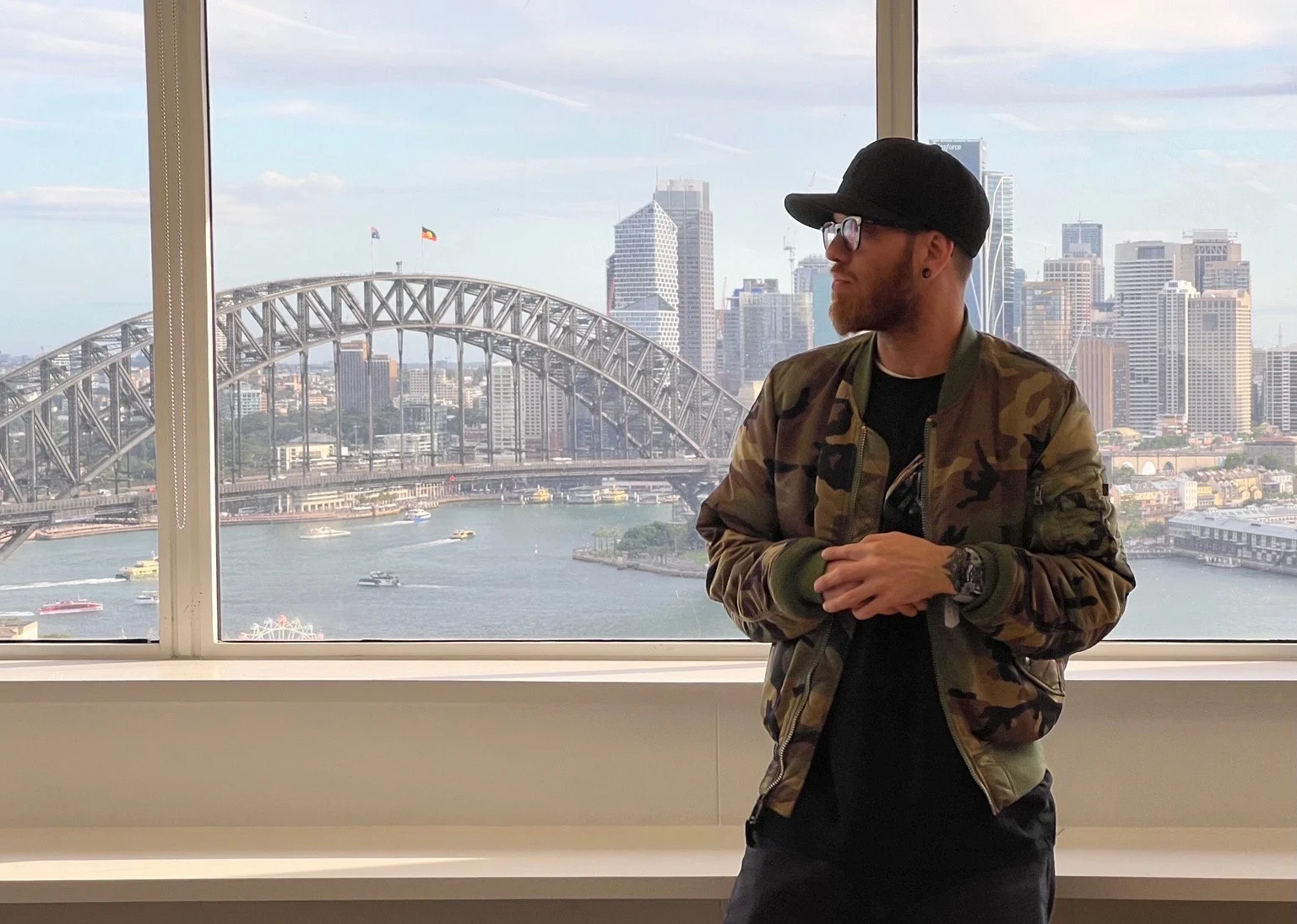 A person in a camouflage jacket and black cap stands at a window with a harbour, arched bridge and city skyline behind.