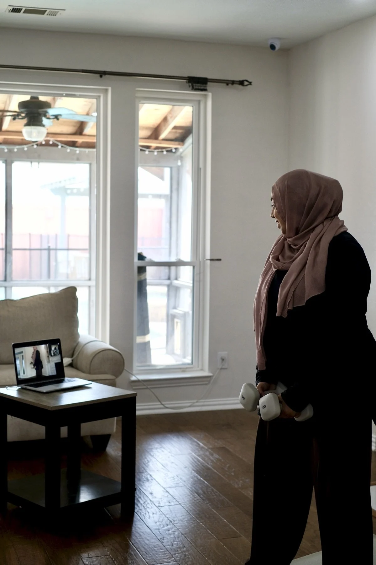 A pregnant Jeem member in hijab holding dumbbells and following a live class on her laptop