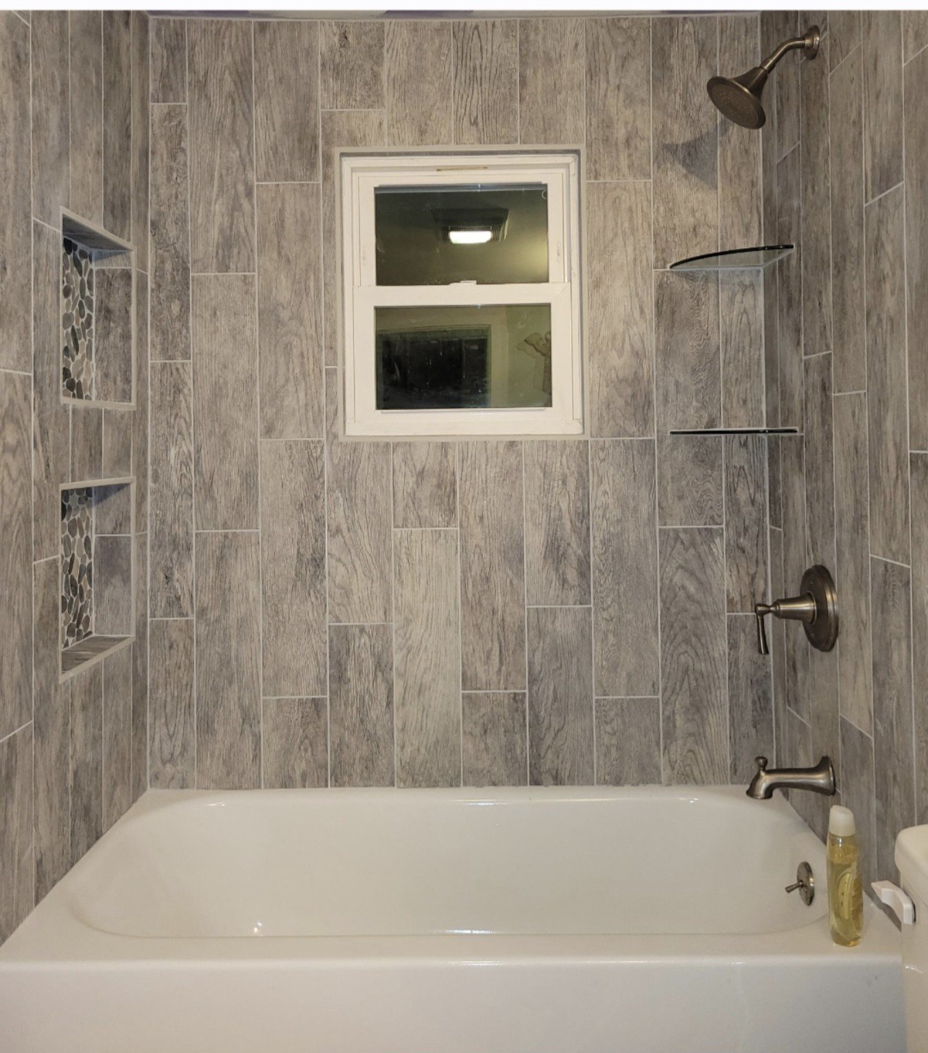 Finished wood-look tile tub surround with a centered window, mosaic niches, corner shelves, and bronze fixtures