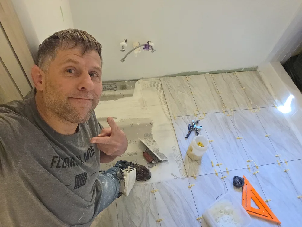 Jesse beside a tile floor installation with leveling clips, exposed mortar, and tile-setting tools visible.