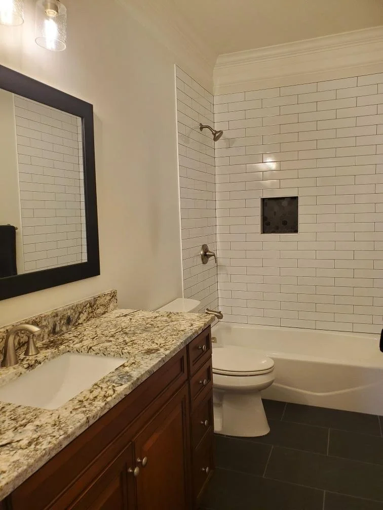 Wood bathroom vanity with a patterned countertop, toilet, and white tiled tub surround.