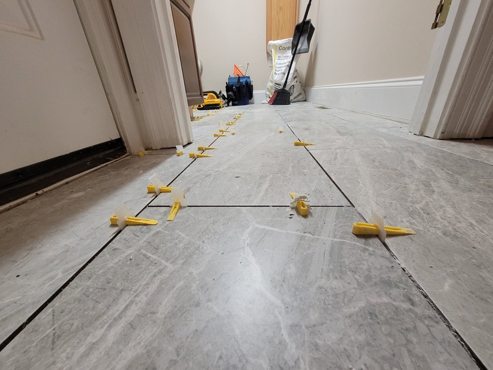 Gray wood-look plank tiles being installed across a bathroom floor with yellow leveling clips along the joints.