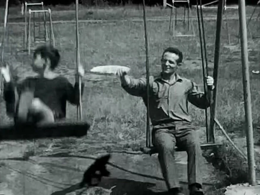 Black-and-white home-movie footage of two people swinging at a playground.