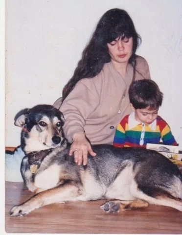 A woman and a child sit behind a large dog indoors.